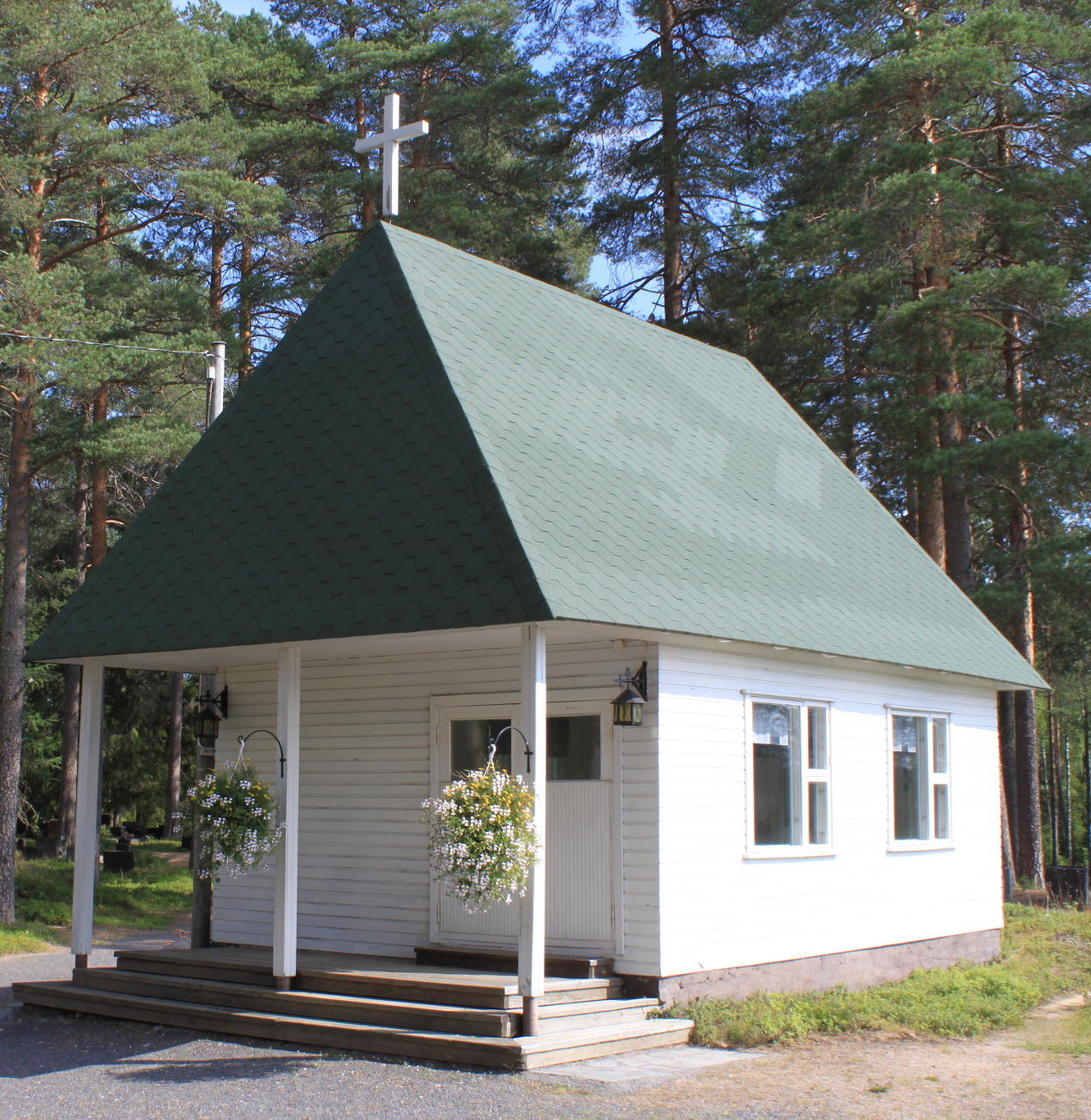 Juorkunan kappeli kesällä: vihreä katto, jossa valkoinen risti. Kukka-amppelit ovella.